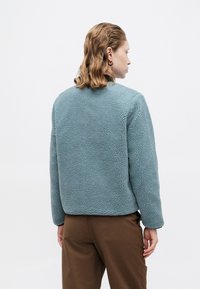 Puha kék fleece dzseki texturált, bolyhos felülettel. Hosszú ujjú, kerek nyakkivágással és egyenes aljjal rendelkezik.