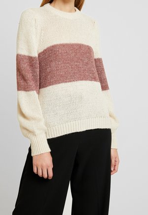 Mujer con un suéter de punto color crema con una amplia franja horizontal color malva a lo largo del pecho y las mangas, combinado con pantalones negros.