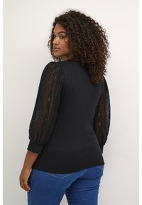 Schwarzes Oberteil mit durchscheinenden Spitzen-Puffärmeln, Rundhalsausschnitt und tailliertem Oberteil, kombiniert mit blauen Jeans. Die Spitze hat ein florales Muster.