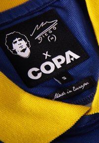 Blauwe en gele polo met kraag, voorzien van een zwart label met witte tekst "COPA" en een handtekening, inclusief een portret van Diego Maradona. Maatlabel erbij.
