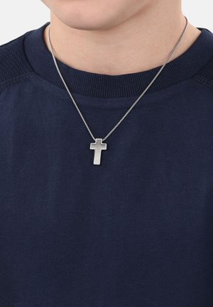 Ciondolo a forma di croce argento su una catena sottile, indossato su una camicia blu navy scura con scollo a costine. Focalizzazione sulla texture e sul design del metallo.