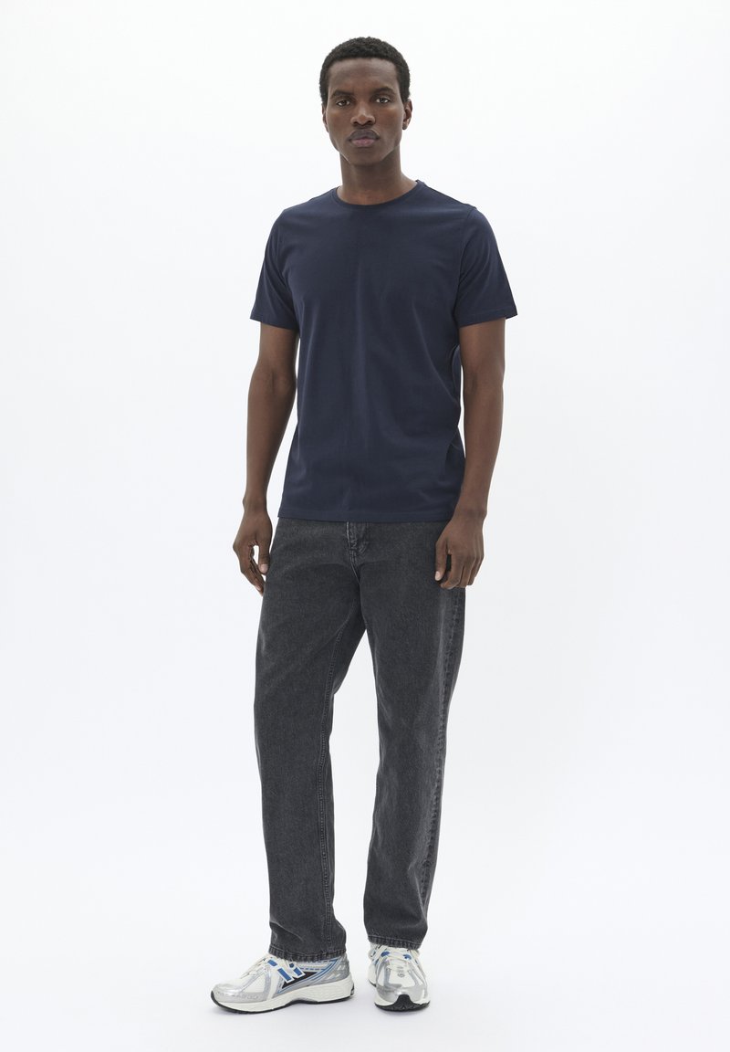 Camiseta de manga corta azul marino combinada con jeans gris oscuro y zapatillas deportivas plateadas, de pie contra un fondo blanco liso.