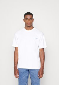 Witte katoenen t-shirt met korte mouwen met een blauw logo-tekst "QUEENS BOROUGH" op de linkerborst, gecombineerd met lichtblauwe denim jeans.