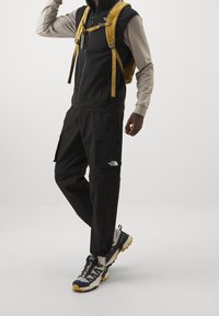 Czarna kamizelka zip-up na lekki longsleeve, spodnie cargo z kieszeniami, oraz wielokolorowe buty turystyczne. Posiada żółte szelki plecaka.