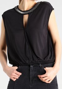 Blouse noire avec un plissement à la taille, présentant un décolleté en trou de serrure orné de détails en perles et des manches courtes. Texture de tissu lisse.