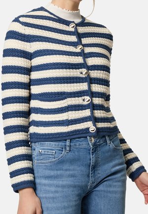 Cardigan en maille rayé bleu et blanc avec des boutons argentés porté sur un col roulé blanc à volants et un jean bleu.