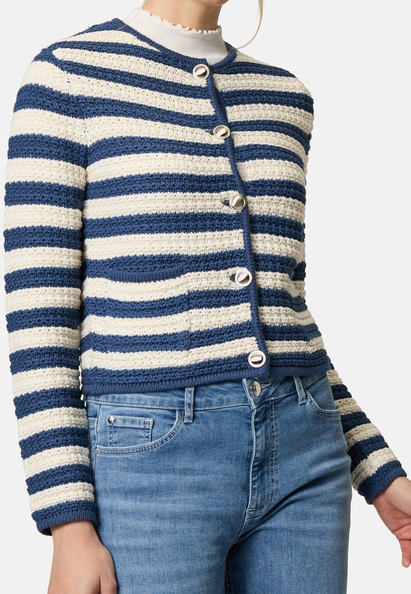 Cardigan en maille rayé bleu et blanc avec des boutons argentés porté sur un col roulé blanc à volants et un jean bleu.
