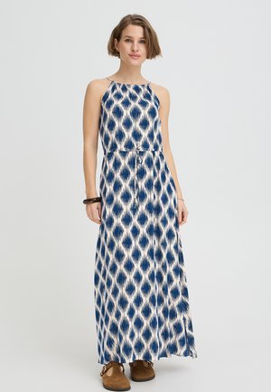 Naine kannab varrukateta sinise ja valge mustriga maxi kleiti, millel on nööriga vöökoht, ja pruune kingi, seistes lihtsa tausta ees.