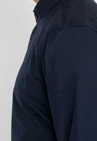 Camisa azul marino de botones con cuello estructurado, fabricada en algodón suave y de corte entallado, que muestra vistas laterales de las costuras y los botones.