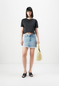 Mulher vestindo uma t-shirt preta cropped, saia de ganga clara desfiada, sandálias de salto pretas, segurando uma bolsa amarelo pálido, de pé contra um fundo branco.