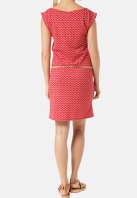 Czerwona sukienka w wzór chevron, z krótkimi falbankowymi rękawami i dopasowanym krojem. Złoty akcent w postaci pasa w talii oraz długość do kolan. Noszona z płaskimi sandałami.