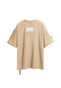 Camiseta de manga corta ribeteada beige con cuello redondo, que presenta una etiqueta blanca en la parte posterior y un lazo de tela en el dobladillo.