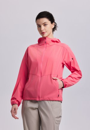 Joven con cabello oscuro que lleva una chaqueta rosa brillante con cremallera y pantalones beige, de pie frente a un fondo liso de color gris claro.