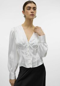 Blusa de satén blanco con un escote en V profundo, torso estructurado, detalles de botones y mangas largas abullonadas, combinada con una falda negra.