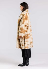 Faux fur-kappa i vitt med bruna fläckar, oversized-design, enkel knappstängning. Bärs över en svart plisserad kjol och spetsiga svarta stövlar.