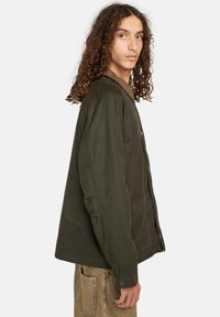 Olivgrüne, knopfrei zu schließende Jacke mit Kragen, zwei vorderen Taschen und einer lässigen Passform. Der Stoff wirkt strukturiert und langlebig.