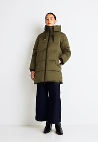 Olivgrön vadderad jacka med huva, med en framsida som har dragkedja, sidofickor och en något längre baksida för extra täckning. Bärs med mörka jeans.