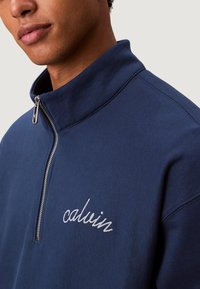 Sudadera azul marino con cuello alto, cierre de media cremallera y detalle bordado "calvin". La tela parece suave con una textura lisa.