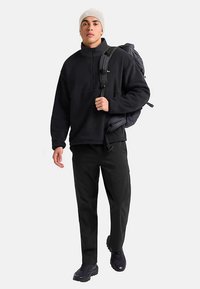 Pull-over en polaire noire avec demi-fermeture éclair, associé à un pantalon noir. Accessoires : un bonnet gris et un sac à dos noir. Sur un fond uni.