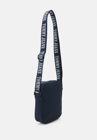 Borsa a tracolla in tessuto blu navy con forma rettangolare e una tracolla audace e marchiata che presenta la scritta "TOMMY JEANS" in bianco. Chiusura con zip.