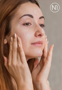 Auftragen eines Gesichtsserums mit einem klaren Gel auf der Haut. Lange braune Haare, natürliches Make-up und metallisch goldener Nagellack an den Händen. Neutraler Hintergrund.