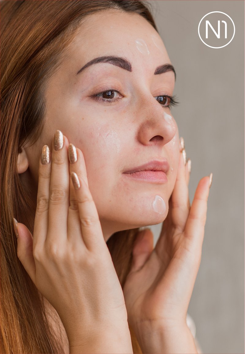 Auftragen eines Gesichtsserums mit einem klaren Gel auf der Haut. Lange braune Haare, natürliches Make-up und metallisch goldener Nagellack an den Händen. Neutraler Hintergrund.