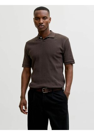 Mees kannab pruuni lühikese varrukaga tekstuuriga polosärki, musti pükse ja pruuni vööd heleda tausta ees.