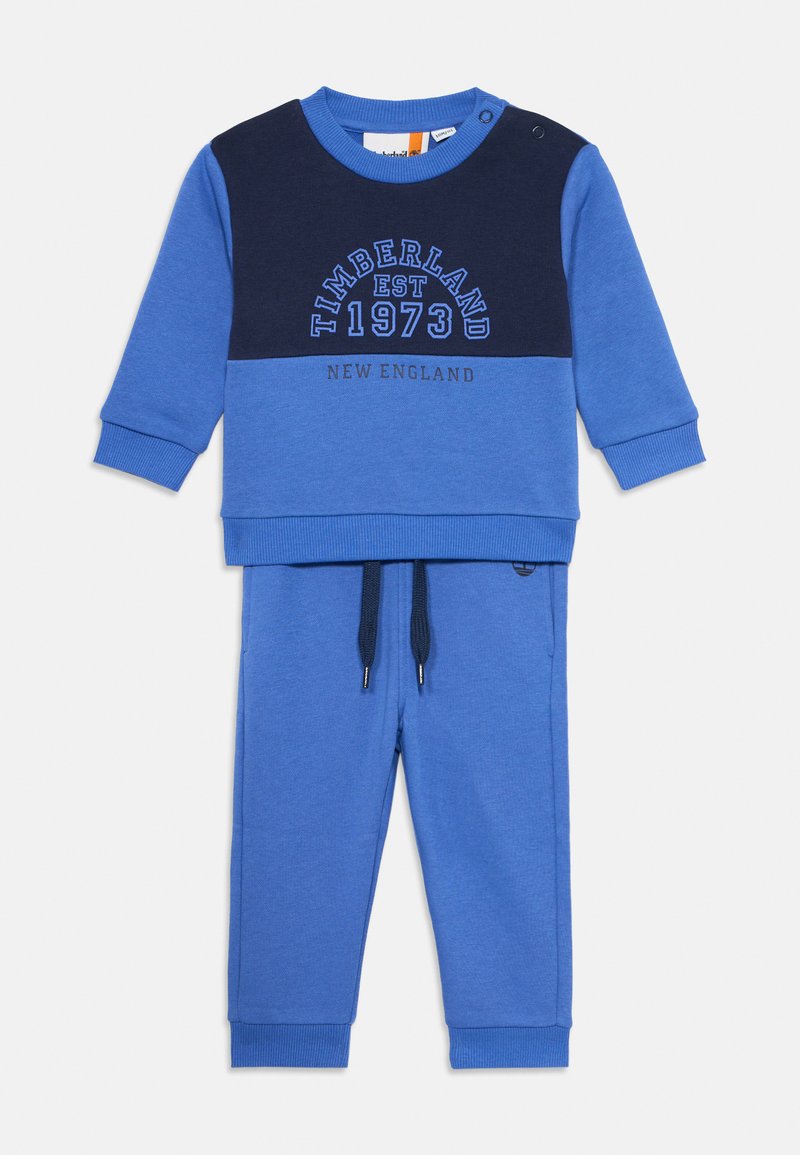 Conjunto de duas peças azul composto por um sweatshirt de gola redonda com painel azul-marinho, logótipo bordado e calças de jogging coordenadas com cordão.