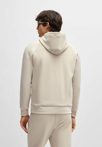 Haut à capuche beige avec une texture douce, doté d'une capuche à cordon, de poignets et d'un ourlet côtelés, et de manches raglan. Porté avec un bas assorti.