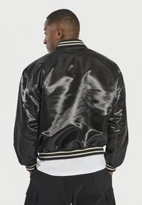 Chaqueta bomber de satén negro con cuello y puños acanalados, que presenta un trim estriado en contraste, se muestra desde la parte posterior con una textura suave.