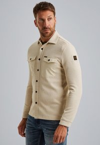 Beżowa koszula z guzikami, z teksturowanym wzorem dzianiny, dwoma kieszeniami na piersi, czarnymi guzikami i naszywką z logo na lewym rękawie.
