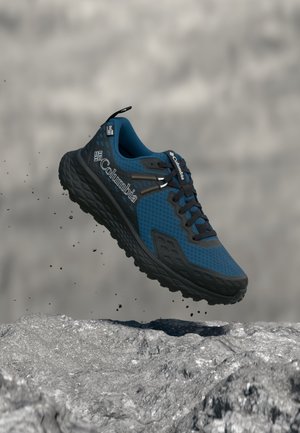 Chaussure de trail Columbia bleue et noire en l’air au-dessus d’une surface rocheuse avec de petites particules de terre qui tombent autour.