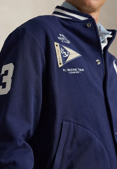 Άνδρας που φορά σκούρο μπλε varsity μπουφάν με ριγέ γιακά, λευκό αριθμό 3 στο μανίκι, ταμπέλες με ναυτικό θέμα και κλείσιμο με κουμπιά τύπου snap.