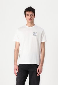 T-shirt en coton blanc avec des manches courtes, col rond, et un petit logo imprimé en gris et noir sur la partie gauche de la poitrine. Porté avec un jean foncé.