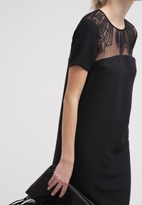 Černé šaty s krátkými rukávy, průhledným vzorovaným výstřihem a rovnou siluetou. V kombinaci s černou třásňovou kabelkou.