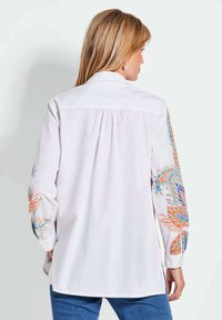 Chemise blanche avec broderies florales colorées sur les manches, col boutonné et manches longues. Ourlet arrondi et texture de tissu légère.