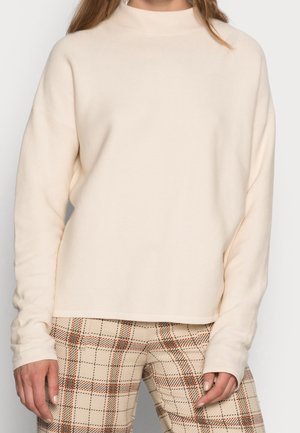Personne portant un pull à manches longues couleur crème et un pantalon beige à carreaux avec des lignes orange et noires, debout les mains relâchées le long du corps.