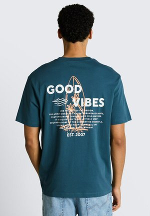 Mann trägt ein türkisfarbenes T-Shirt mit dem Schriftzug "GOOD VIBES" und einer Surfbrett-Grafik, kombiniert mit hellblauen Jeans, den Rücken zur Kamera gewandt.
