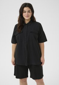 Chemise noire à boutons avec des manches courtes, deux poches avant et coupe ample. Associée avec un short noir assorti, fabriqué en tissu léger.