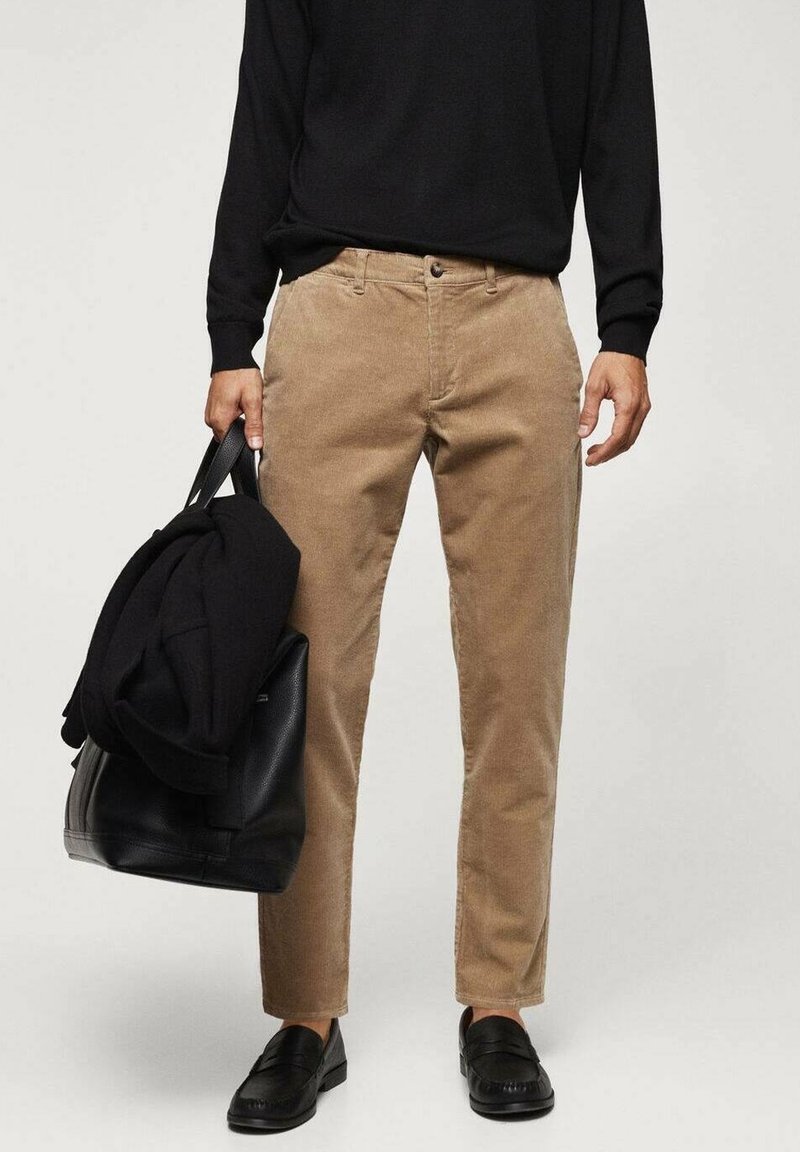 Homme portant un pantalon en velours côtelé beige et des mocassins noirs, tenant un sac noir avec une veste noire pliée dessus.