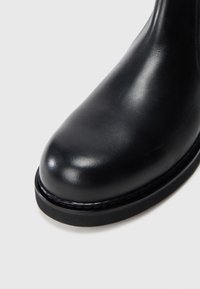 Bota de cuero negra hasta el tobillo con punta redonda, textura suave y un detalle de costura mínimo. La suela es plana con un ligero borde para mayor agarre.