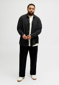 Mees seisab valge tausta taustal, jalas valged tossud, seljas lõtv must püks, valge särk ja lahtise nööbiga must ruuduline särk.