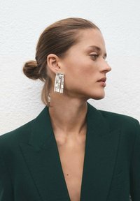 Kryształowe kolczyki z żyrandolem o prostokątnym kształcie, z przezroczystymi kamieniami ułożonymi kaskadowo. Noszone z ciemnozielonym żakietem.