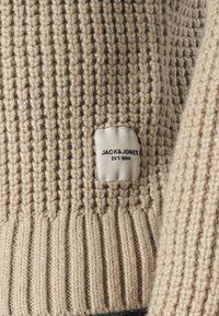 Tessuto di maglione beige lavorato a maglia con un motivo testurizzato, caratterizzato da un'etichetta bianca con la scritta "JACK & JONES EST. 1990" ricamata in nero.