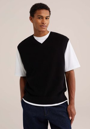 Ung mand iført en sort ærmeløs sweater over en hvid T-shirt og mørkeblå jeans, stående foran en ensfarvet beige baggrund.