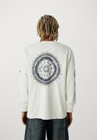 Tricou alb cu mânecă lungă, având un design grafic circular negru pe spate, cu modele și simboluri complexe. Țesătură din bumbac moale.