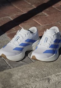 Coppia di scarpe da corsa bianche con strisce blu e testo "ADIZERO", posizionate su pavimentazione in pietra al sole con ombre.