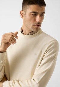 Maglione color crema con trama testurizzata, girocollo e polsini a coste, con un logo discreto sul petto e una camicia con colletto sotto.
