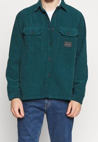 Tealfärgad kordflöjelskjorta med knapp-up front, två bröstfickor och en etikett. Har långa ärmar och en platt krage.