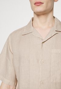 Beige linnen shirt met een ontspannen kraag, korte mouwen, een borstzak en twee knopen. De textuur van de stof geeft een lichte, ademende uitstraling.